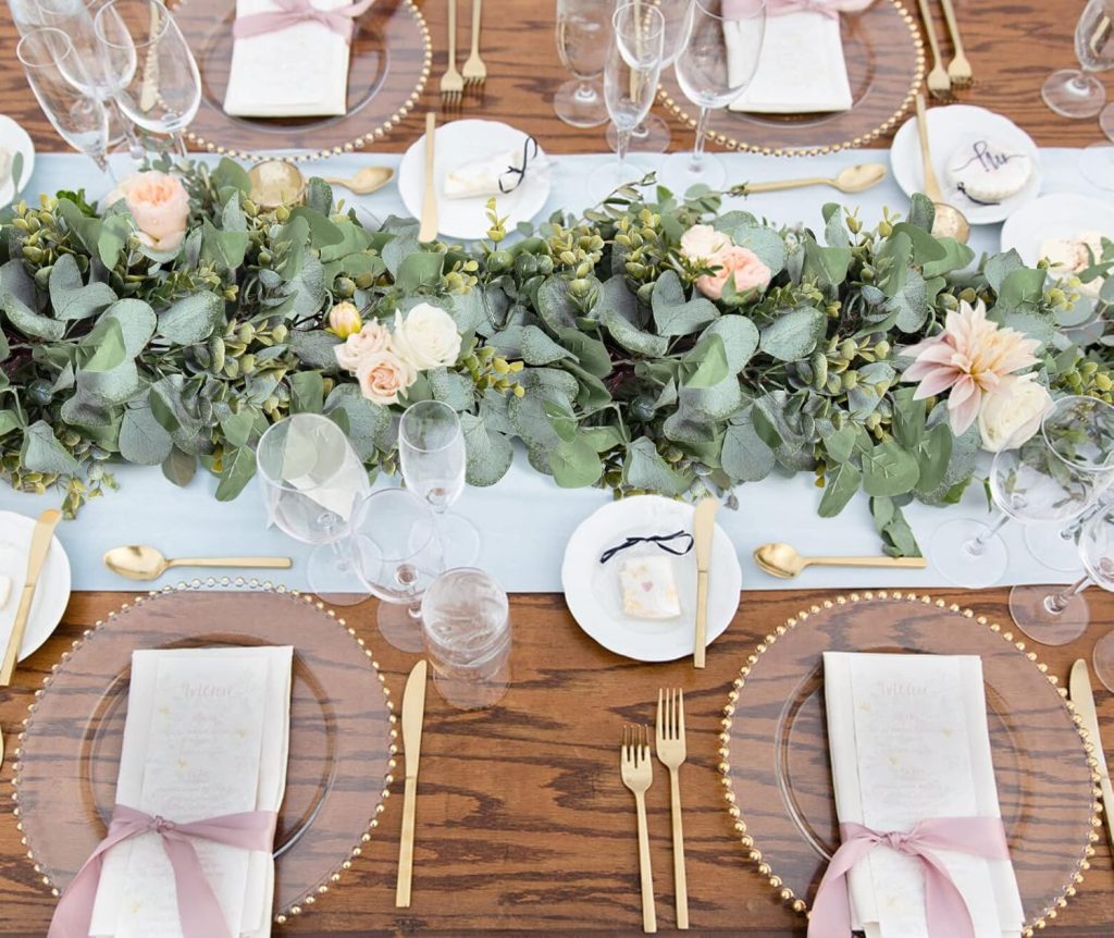 Elegant wedding reception table setting with a eucalyptus and blush flower garland centerpiece, gold cutlery, glass beaded charger plates, and dusty rose napkin ribbons on a rustic wood table.