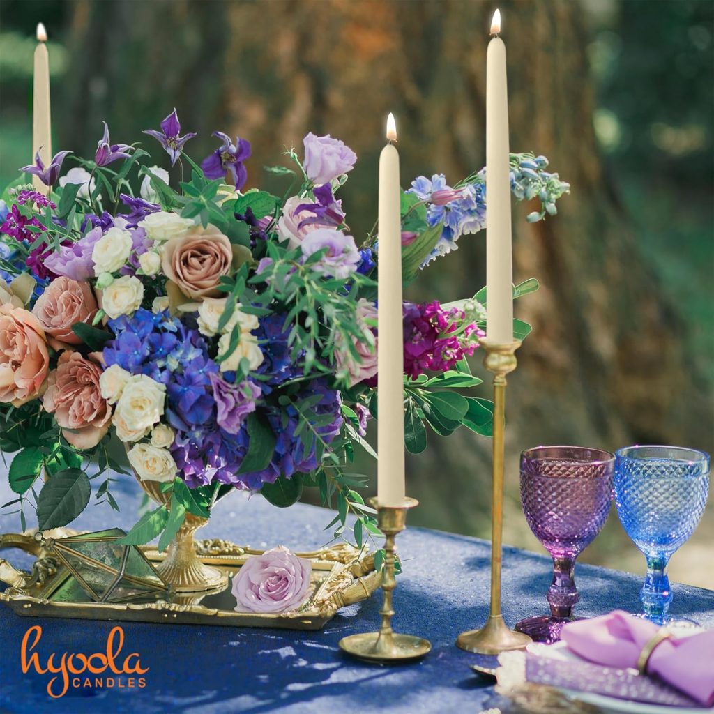 A close-up table setting features an elegant floral centerpiece with lush purple, blue, and pink flowers like delphiniums and roses on a blue tablecloth. Three tall beige taper candles in gold holders flank the arrangement, accompanied by two purple and blue stemmed glasses and folded purple napkins on gold-rimmed plates. The Hyola Candles logo appears in the corner, evoking a romantic outdoor ambiance with a blurred tree in the background.