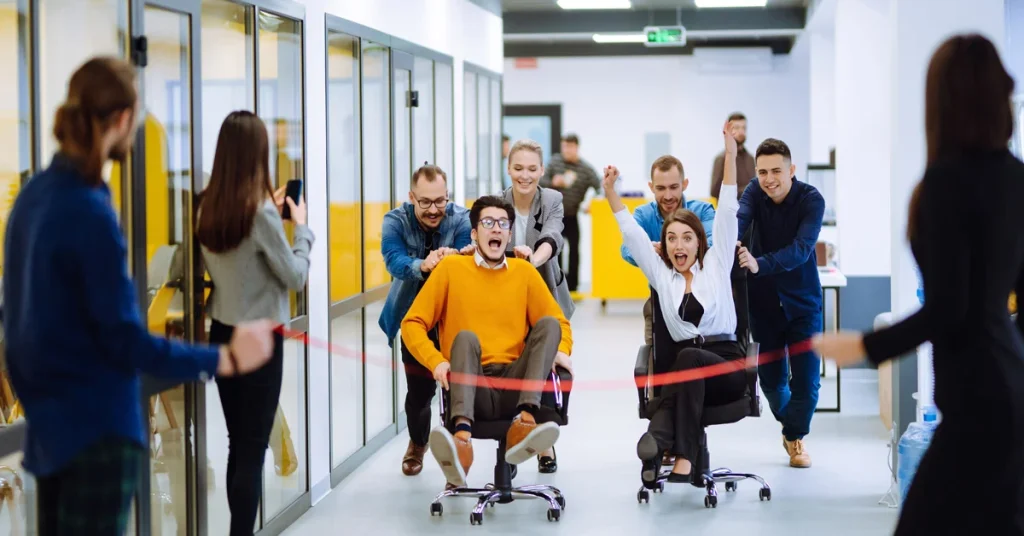 

Coworkers laughing and racing office chairs down a hallway crossing a finish line, a hilarious and memorable office birthday party game idea for the workplace.