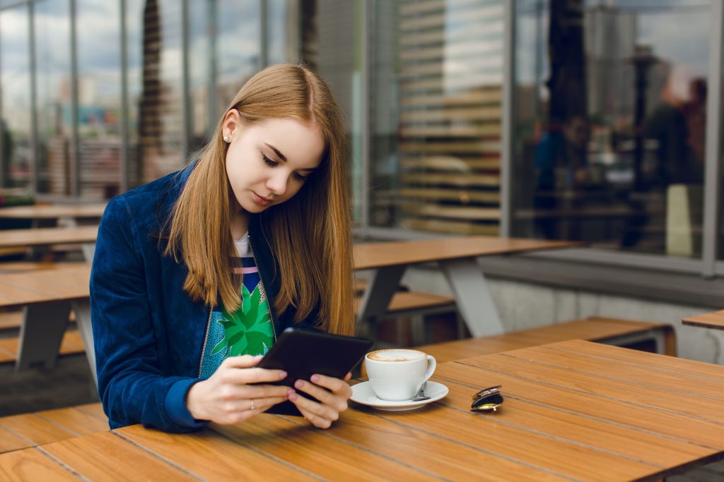 Young woman using a tablet at an outdoor café table with a cup of coffee.