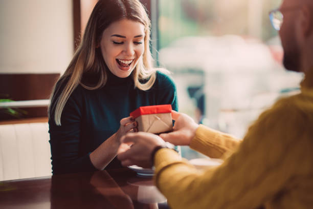 Excited young woman receiving a surprise gift box with red ribbon from partner at cafe — heartwarming winter blues care package moment that brightens a friend's day"